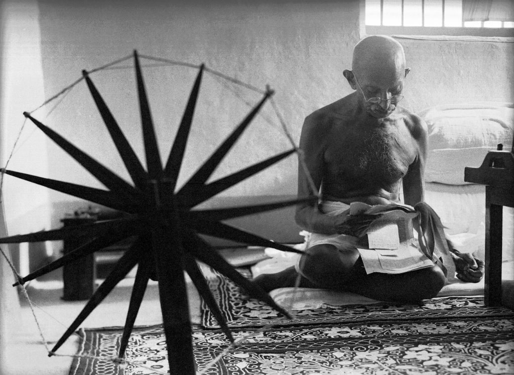 black and white photo of Indian leader Mohandas Gandhi reading next to a spinning wheel at home in India, 1946